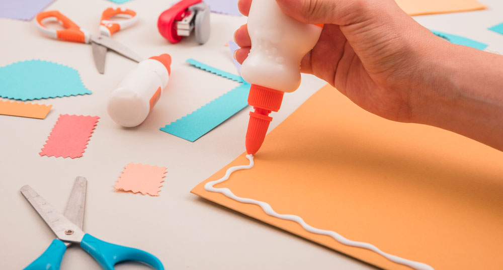 human-hand-applying-white-glue-on-orange-paper-with-scissor-and-stapler_web